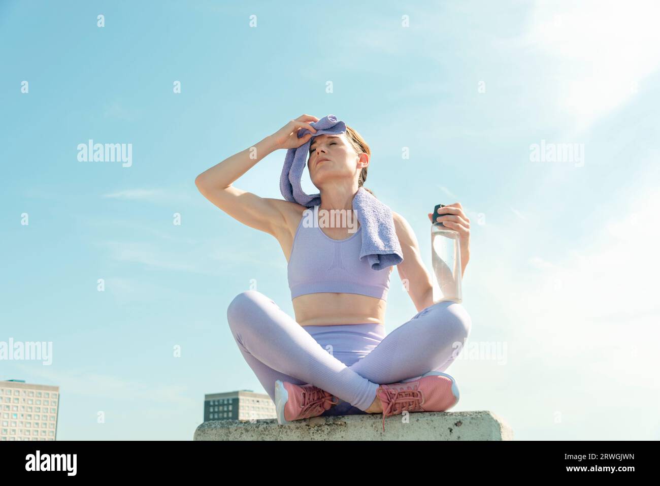 sporty runner sitting wiping head with a towel holding a glass water bottle, urban setting Stock Photo