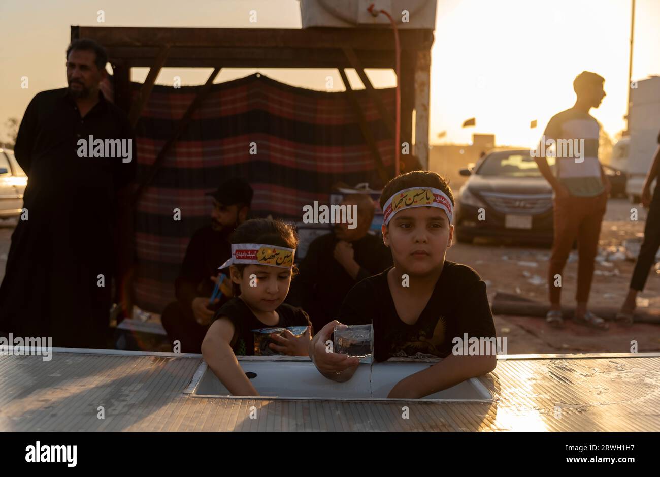 Iraqi kids distribute water among Shia Muslim pilgrims marching from Najaf towards Shrine city of Karbala. Arbaeen is an annual pilgrimage observed by Shia Muslims. Pilgrims walk for about 20 days from cities in Iraq and Iran to the holy city of Karbala, where they commemorate the death of Imam Hussein, the grandson of Prophet Muhammad. Imam Hussein was killed in battle in 680 AD in a conflict against the Caliph Yazid. On the 40th day of mourning for Hussein, the pilgrims arrive in Karbala to pay tribute at his shrine, and millions gather for this final day of Arbaeen. Along the route, volunte Stock Photo