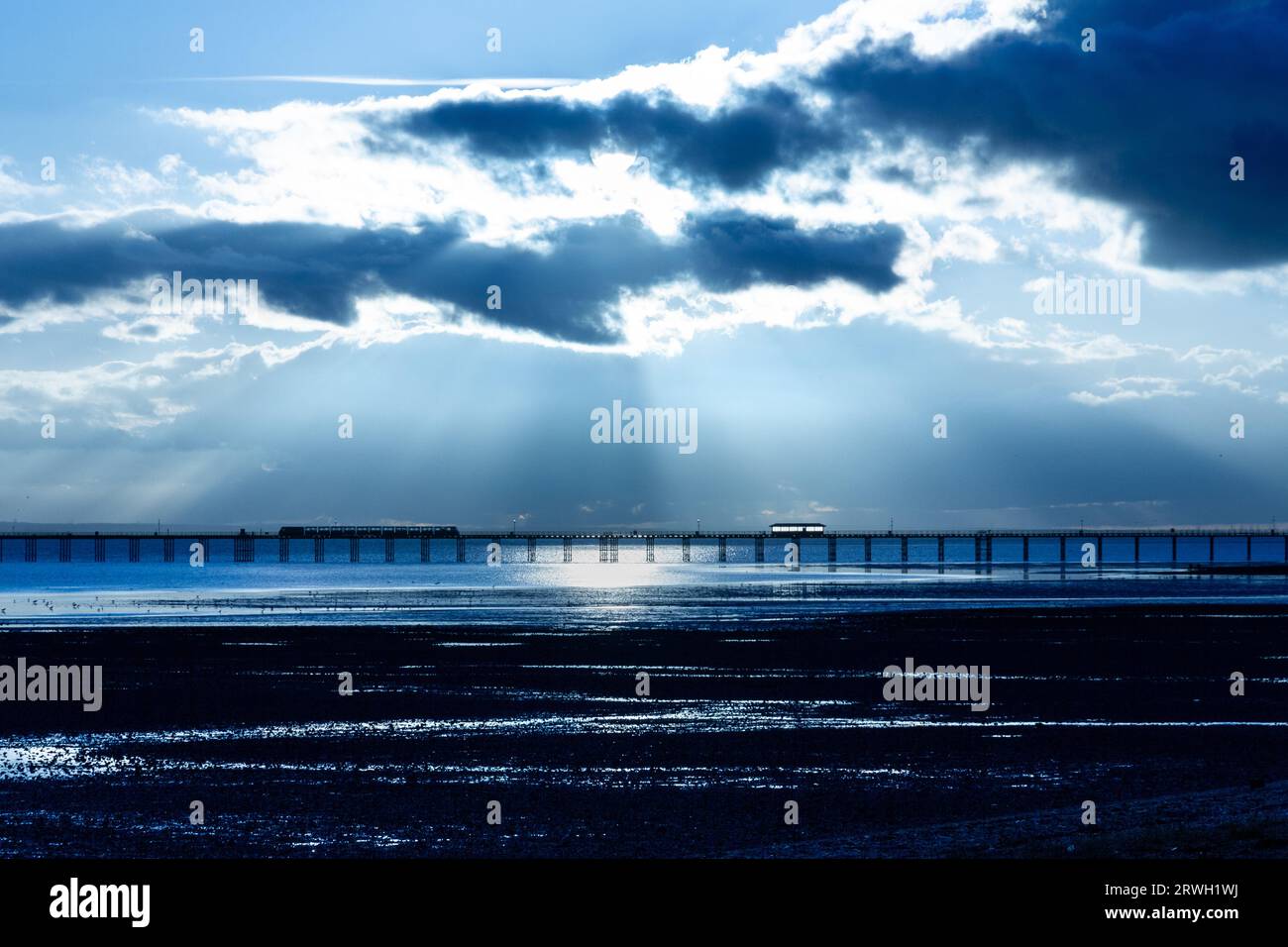A distant view of Southend Pier at dusk with rays of light breaking through the clouds. The tide is out. Stock Photo