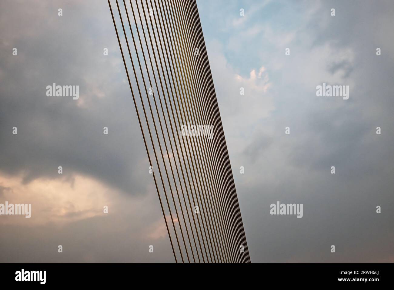 A suspension bridge cable, composed of smaller cables, extends diagonally against a cloudy sky. Stock Photo
