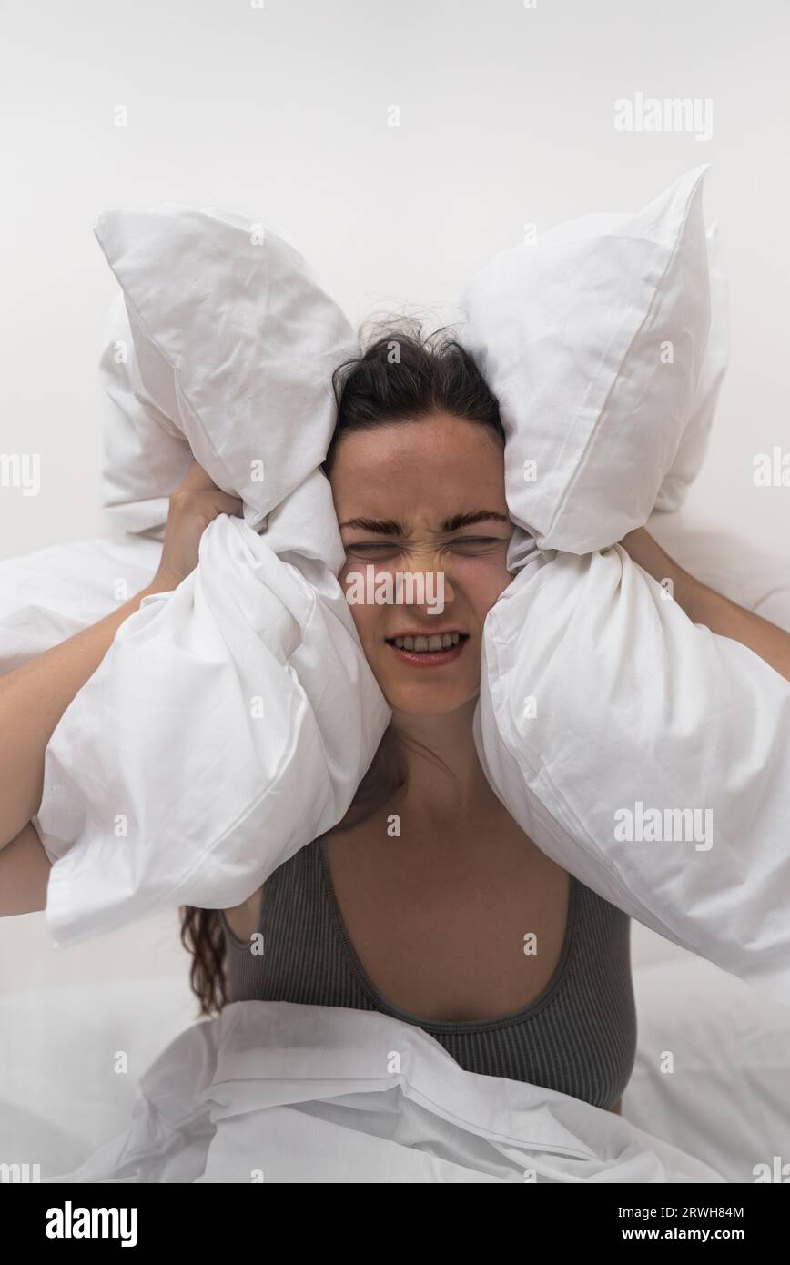 woman covers her ears with pillows, unable to sleep due to noisy neighbors, suffering from