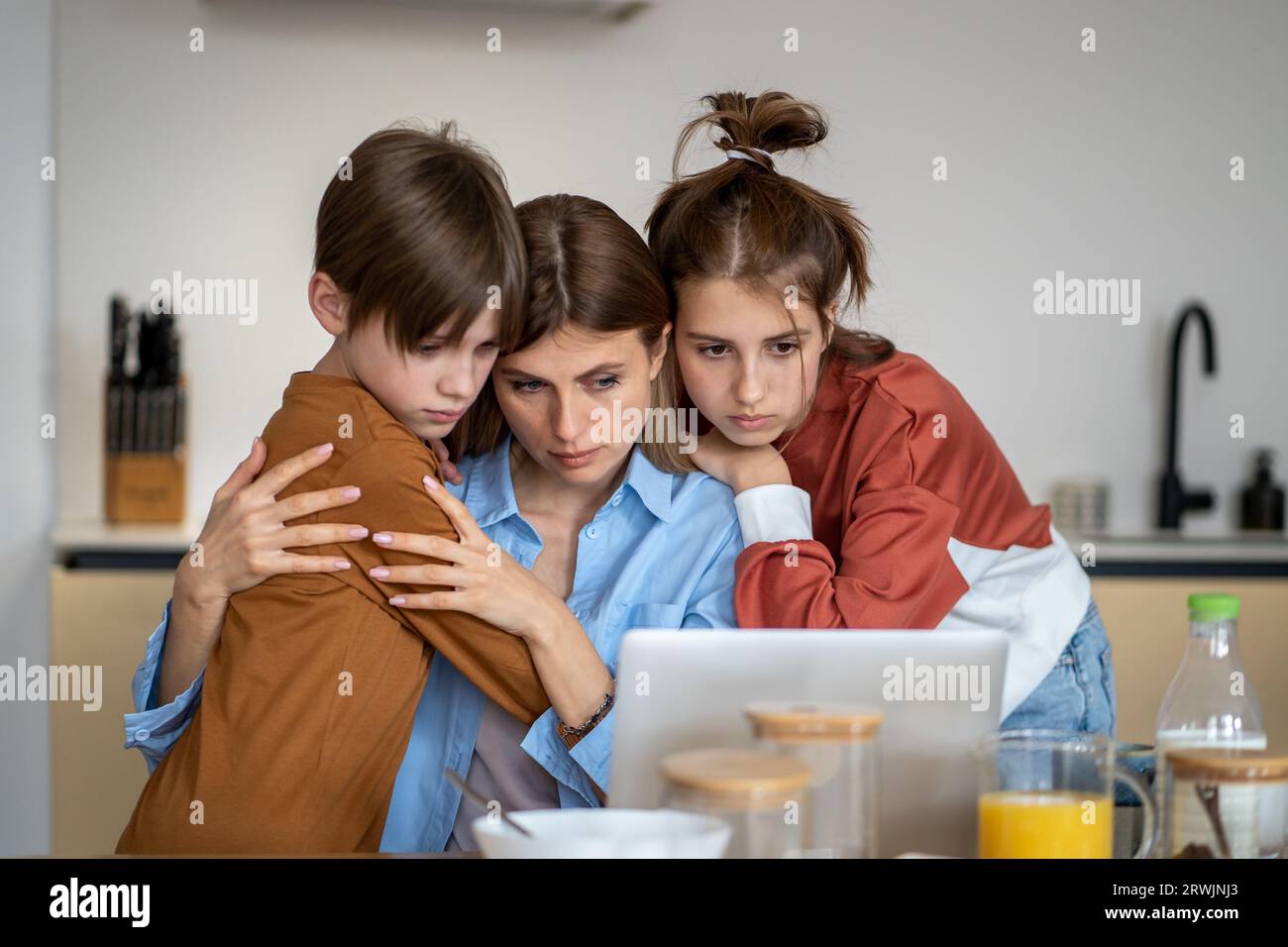 Teenagers children support mother in kitchen looking at laptop screen with bad news from work.  Stock Photo