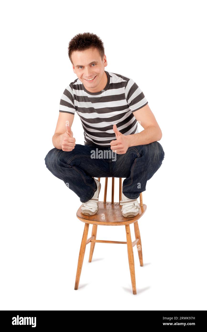 Smiling handsome young man sitting on chair gesturing ok sign isolated against white background Stock Photo
