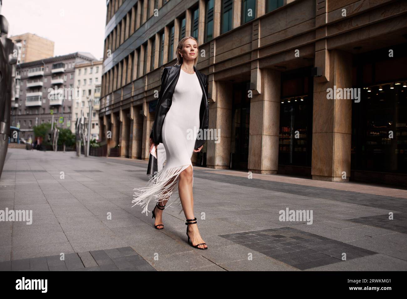 Full length fashionable blonde woman walking city street in chic attire with building in background. Young female model wears white fringed dress, bla Stock Photo