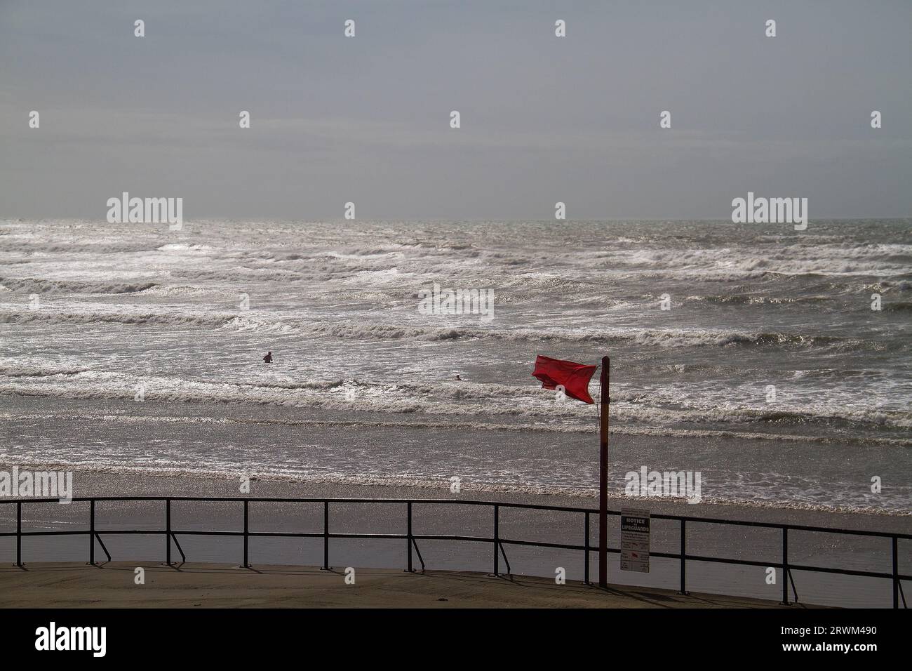 Two people in the distance in a rough sea, ignoring a red flag warning of danger, dark sea reflecting last sunlight Stock Photo