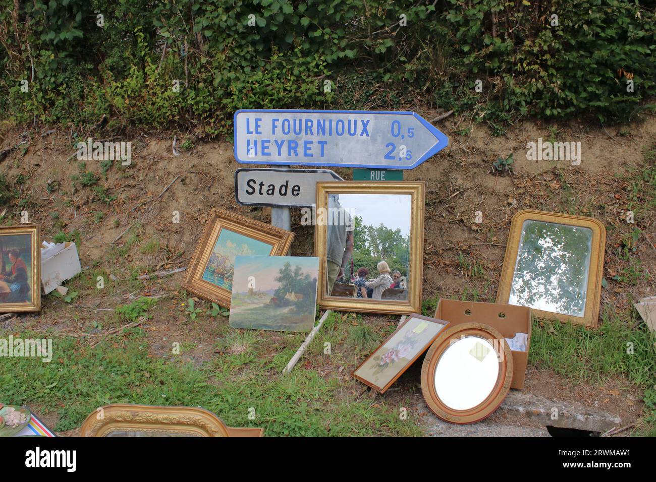 View of paintings and mirrors set against a road sign located at a summer brocante at Chéniers in rural central France. Stock Photo