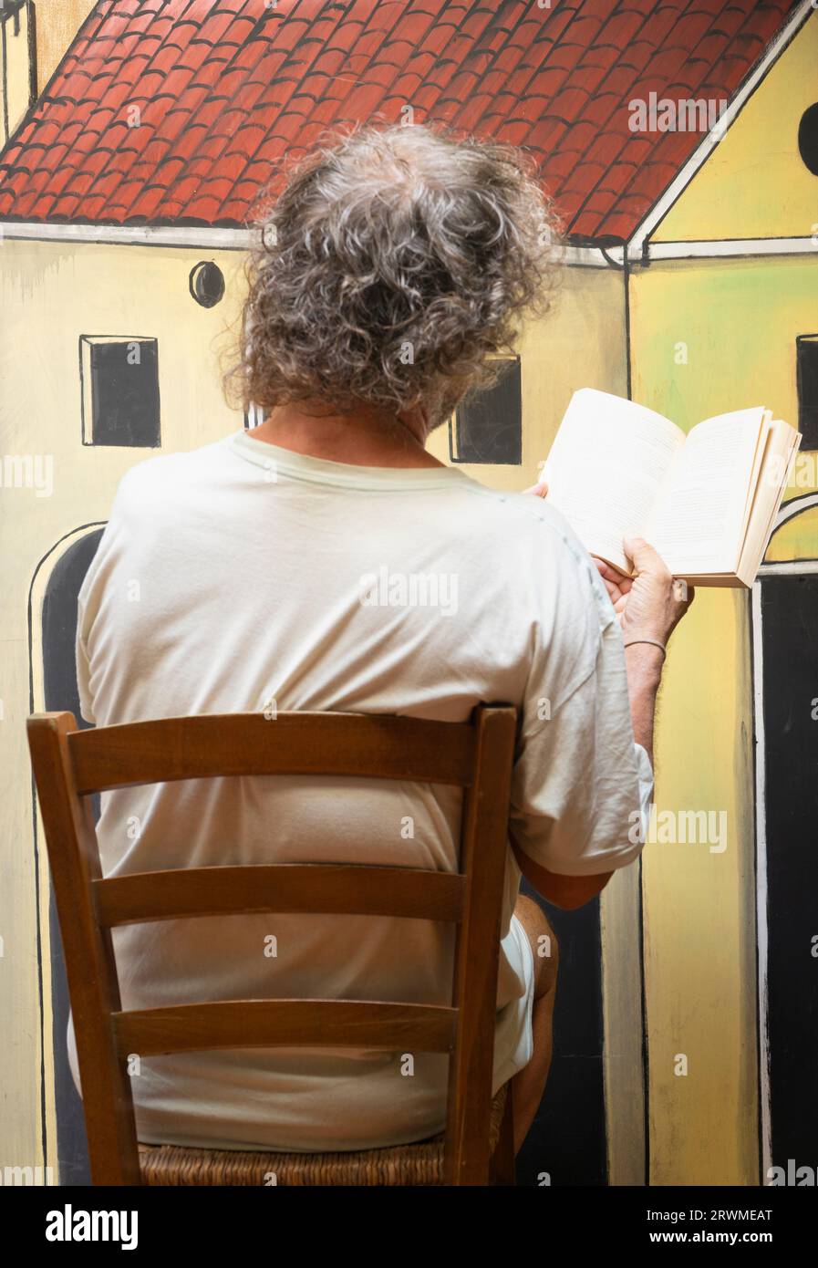 A man from behind sitting in a wooden chair while reading a script in front of a cardboard theater scenery. Stock Photo