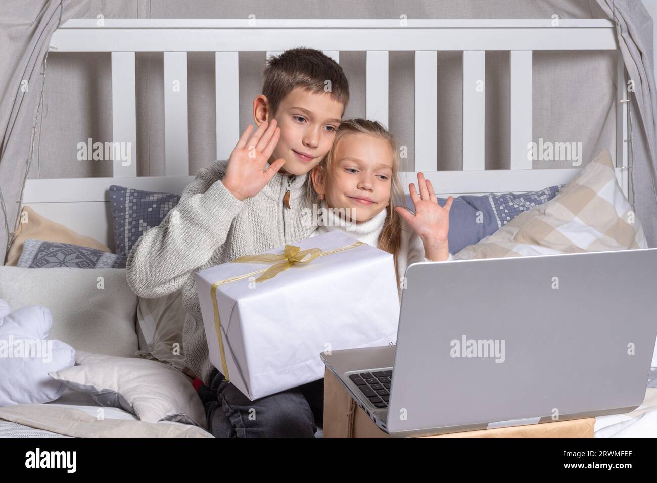 Preschool children holding gift box chatting via video link via laptop and waving hand greeting relatives or friends at home. Boy and girl brother and Stock Photo