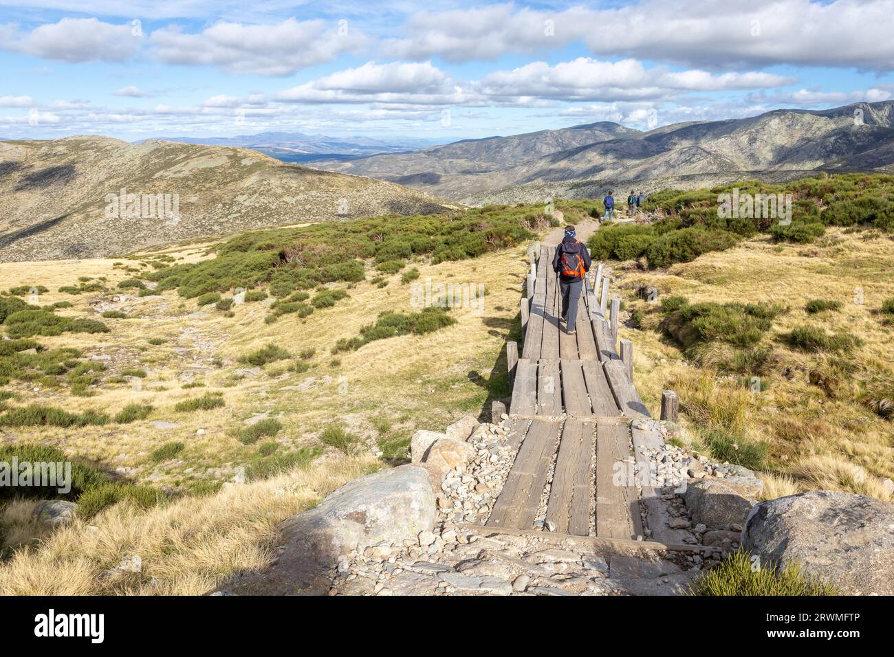 Tourists hiking on wooden trail to the Laguna Grande de Gredos lake from the Plataforma de Gredos in Sierra de Gredos mountains, dry brown grass. Stock Photo
