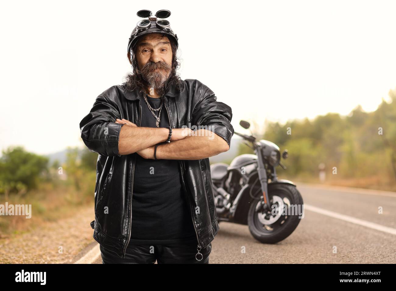 Mature bearded biker with a helmet and leather jacket standing in front of a chopper on the road Stock Photo