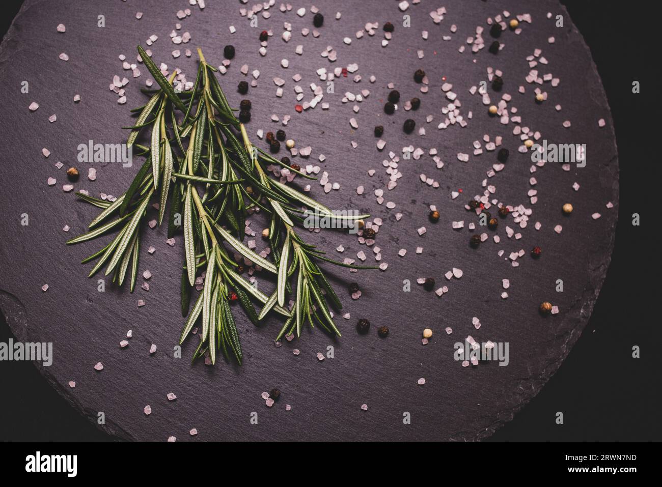 close-up bouquet of rosemary green plants on the table on a black granite plate, scattered Himalayan pink salt and black peppercorns Stock Photo