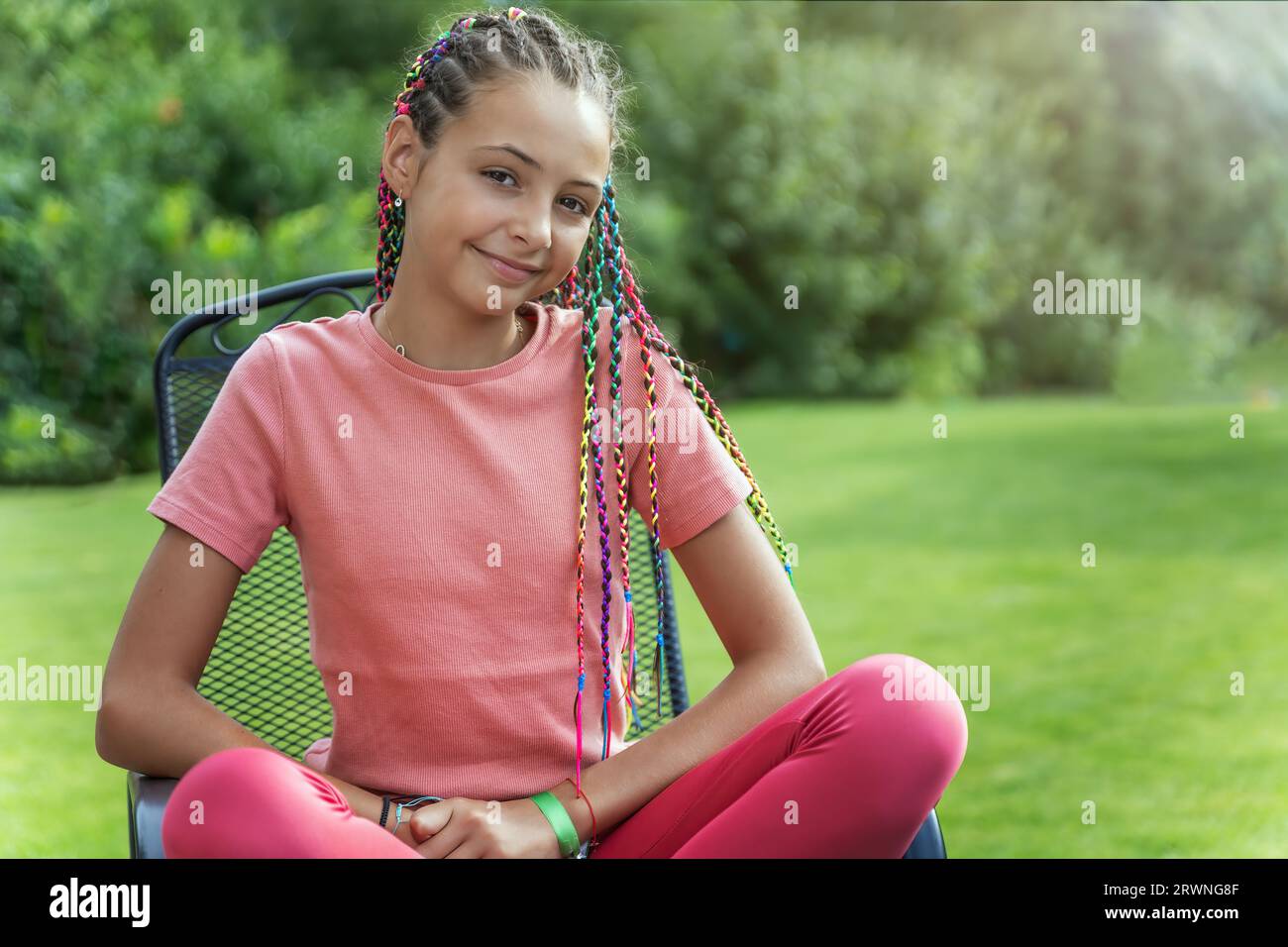 Portrait of smiling young girl with colorful braids in her hair sitting on a chair outside. Horizontally. Stock Photo