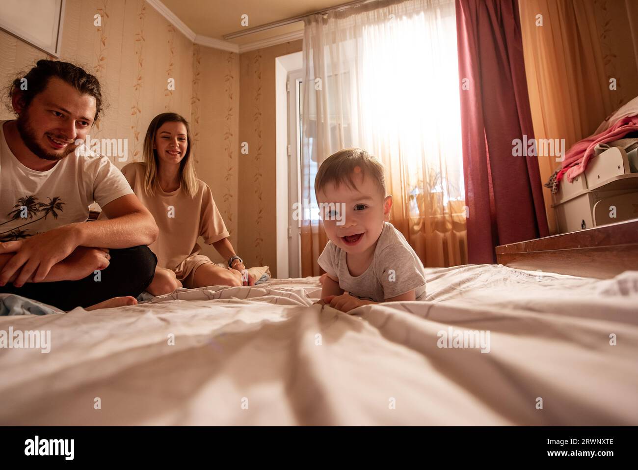 Baby boy lies on stomach on bed, smiling at camera. Family out of focus from behind. Out of focus, father and mother are laughing from behind, looking Stock Photo