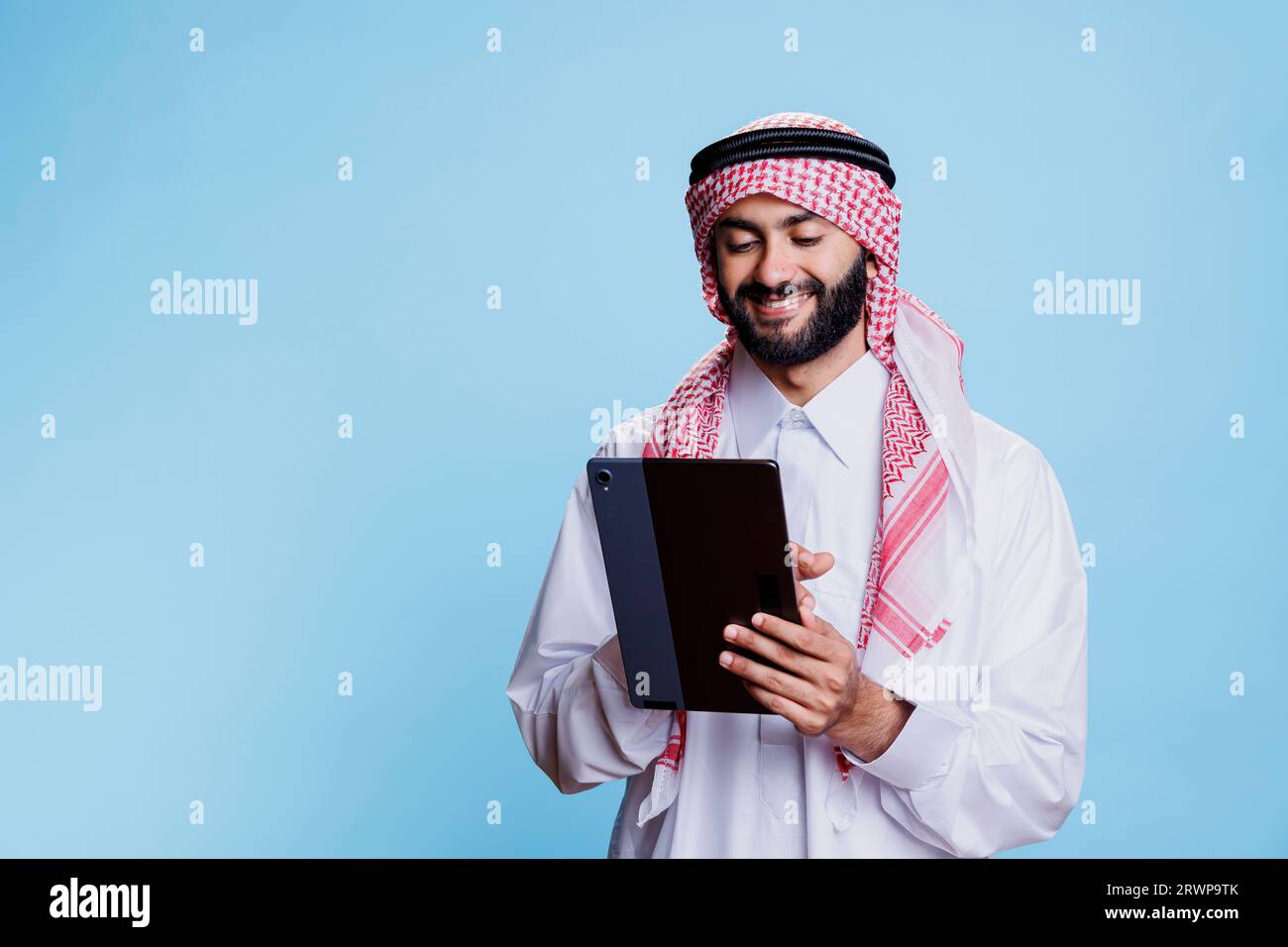 Smiling man wearing muslim headdress and thobe holding digital tablet and tapping on touchscreen. Arab person scrolling internet page on portable gadget with cheerful expression Stock Photo