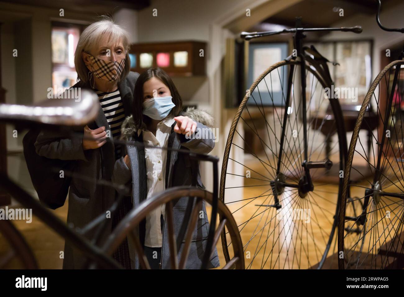 Tween schoolgirl and elderly female tutor in face masks viewing vintage bicycle in museum Stock Photo