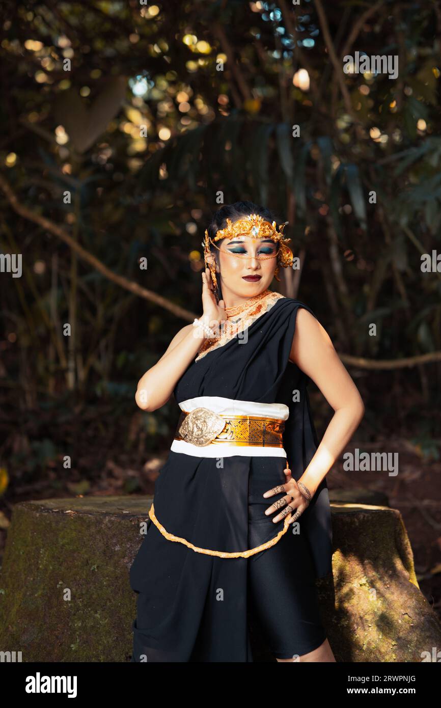 Asian woman standing and posing with her hand in a black costume while wearing a golden crown and golden belt on her body inside the forest Stock Photo