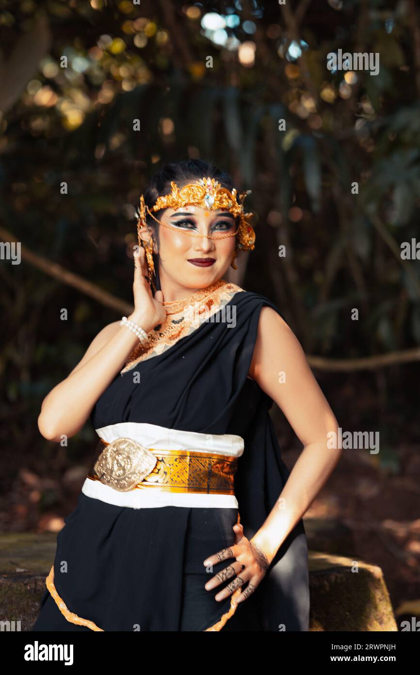 Asian woman standing and posing with her hand in a black costume while wearing a golden crown and golden belt on her body inside the forest Stock Photo