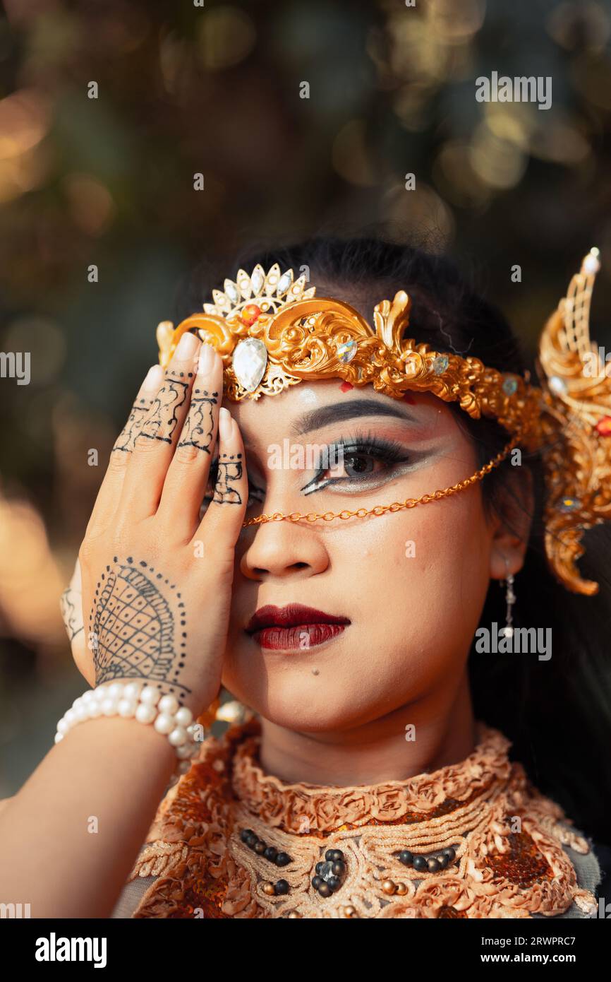 Asian woman covering her face with hands full of tattoos while wearing makeup and a golden headpiece inside the village Stock Photo