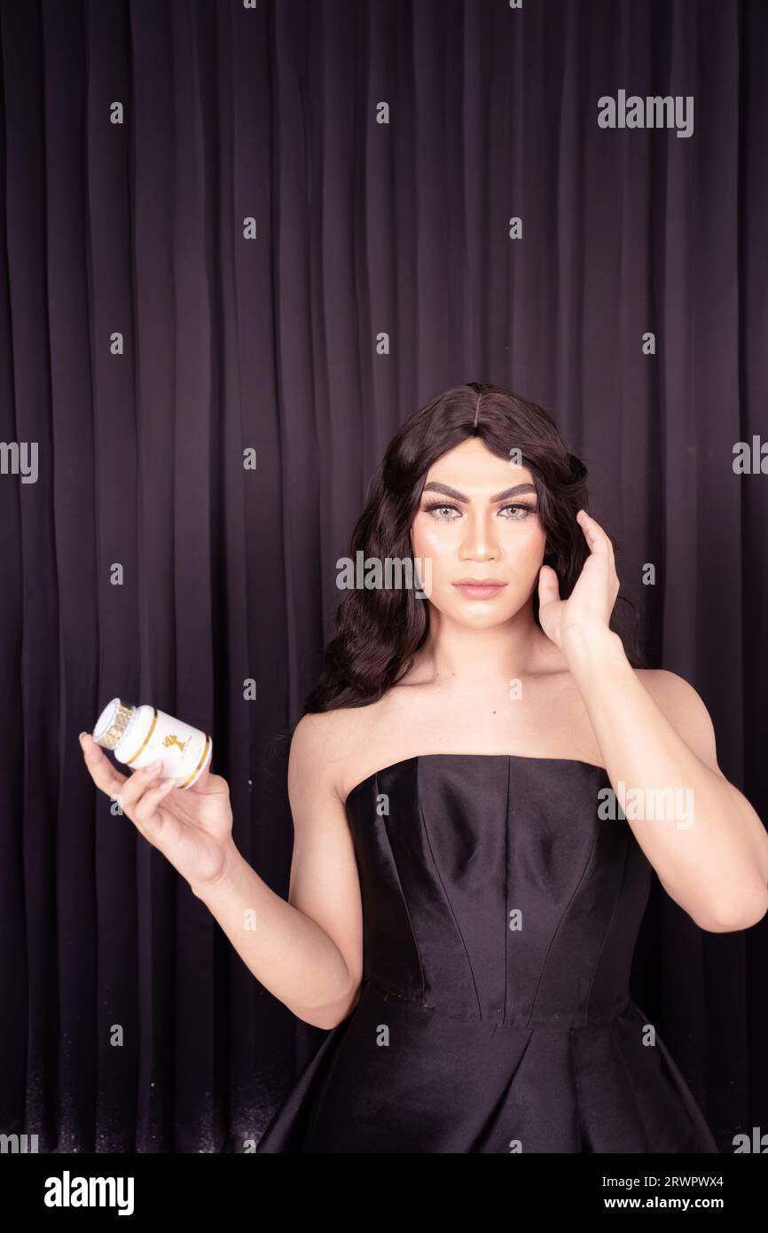 Beautiful Woman holding skincare in her hand while wearing a black dress with black hair in front of the black curtain Stock Photo