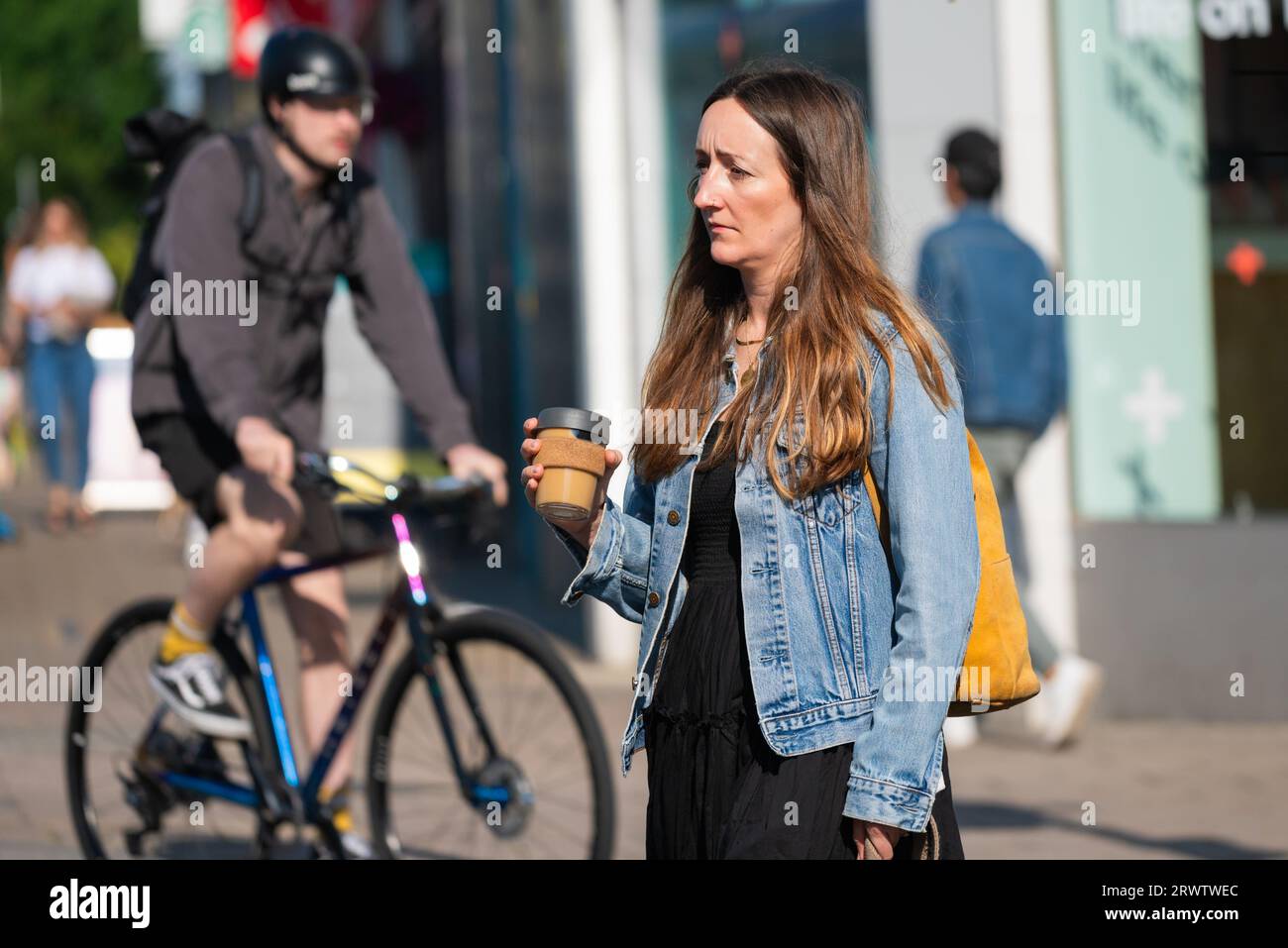 Young to middle aged lady walking across a road carrying a cardboard cup of coffee or another hot beverage, with cyclist in the background. Stock Photo