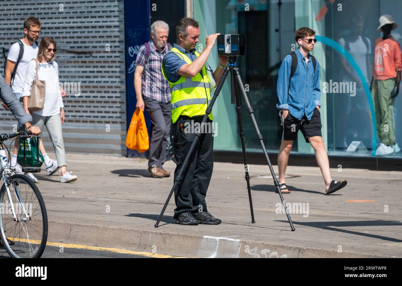 A surveyor or technician in a high-visibility yellow vest operating a 3D laser scanner mounted on a tripod on a busy UK street. Stock Photo