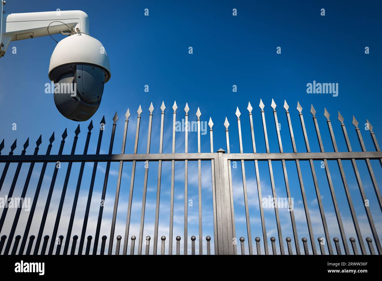 Modern security camera monitoring the entrance to a gate of a private property. Wrought iron gate with sharp points on blue sky and copy space. Stock Photo
