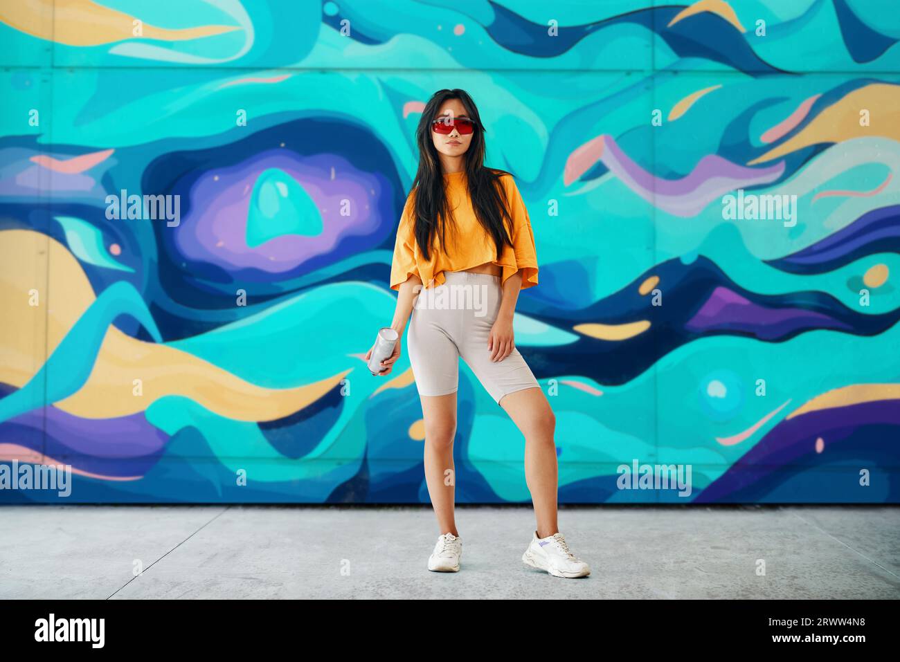 Young female street artist with spray paint can in hand looking to camera over her graffiti wall paintings background. Street art concept. Full length Stock Photo