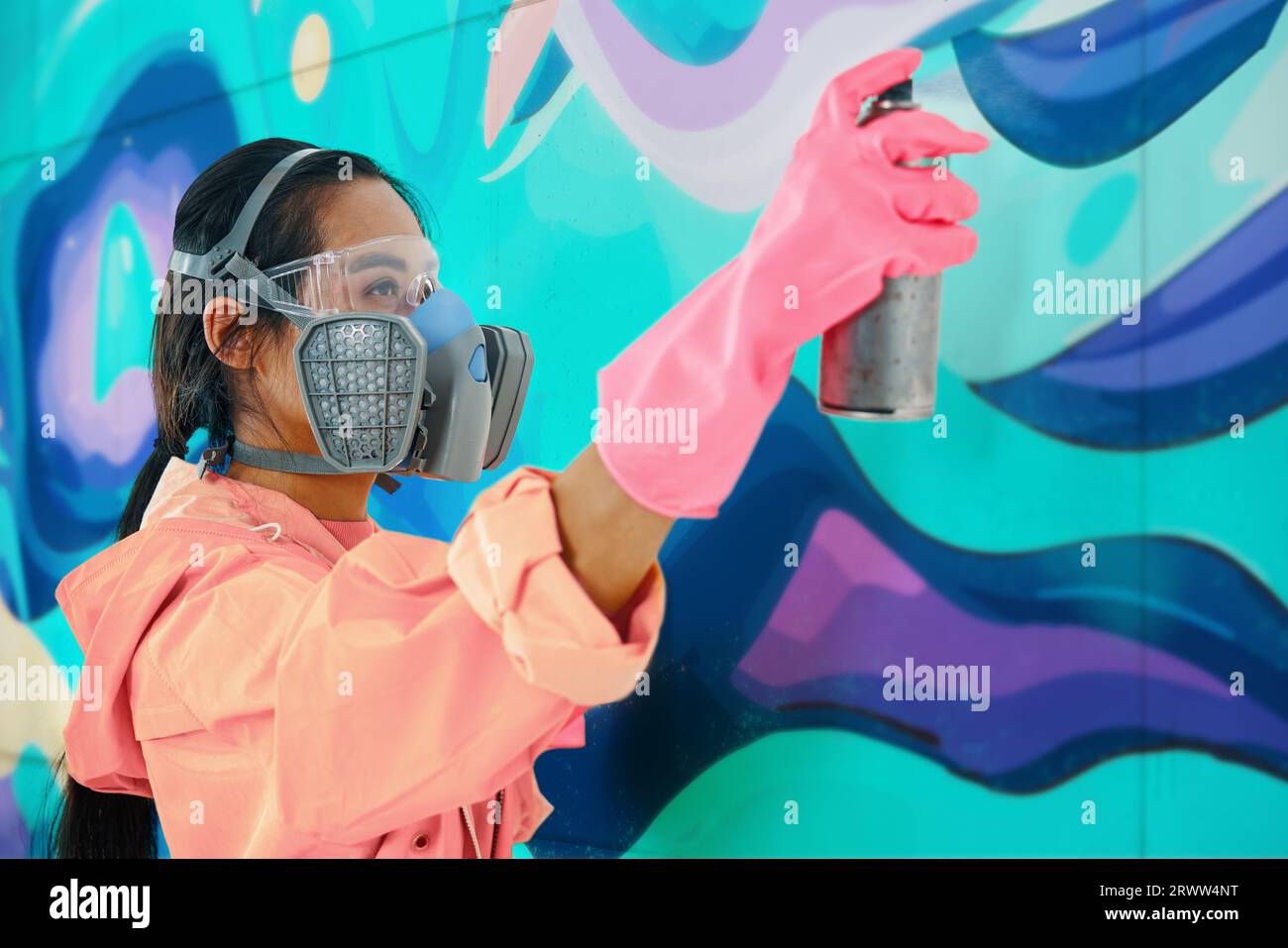 Female street artist painter in respirator mask painting colorful graffiti on wall Modern art, urban concept. Stock Photo
