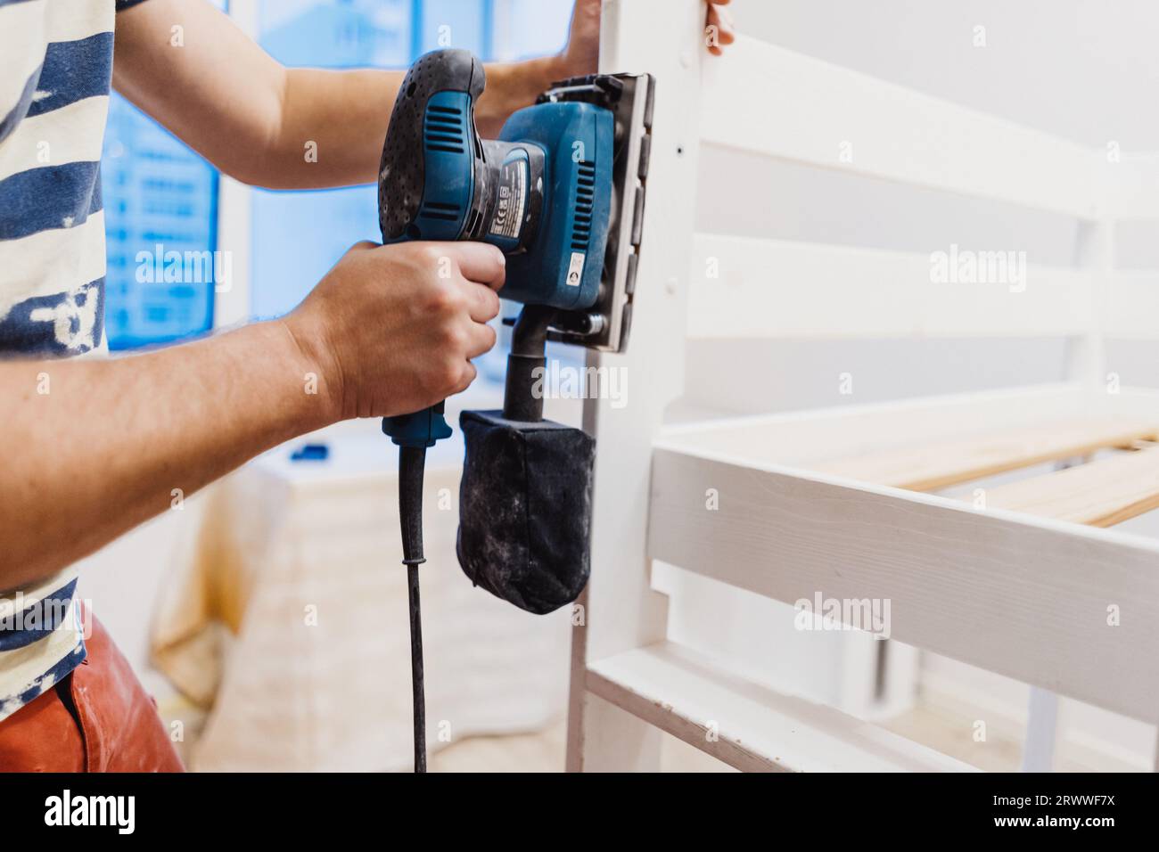 unrecognizable man rubs wooden child bed with power sander or grinder