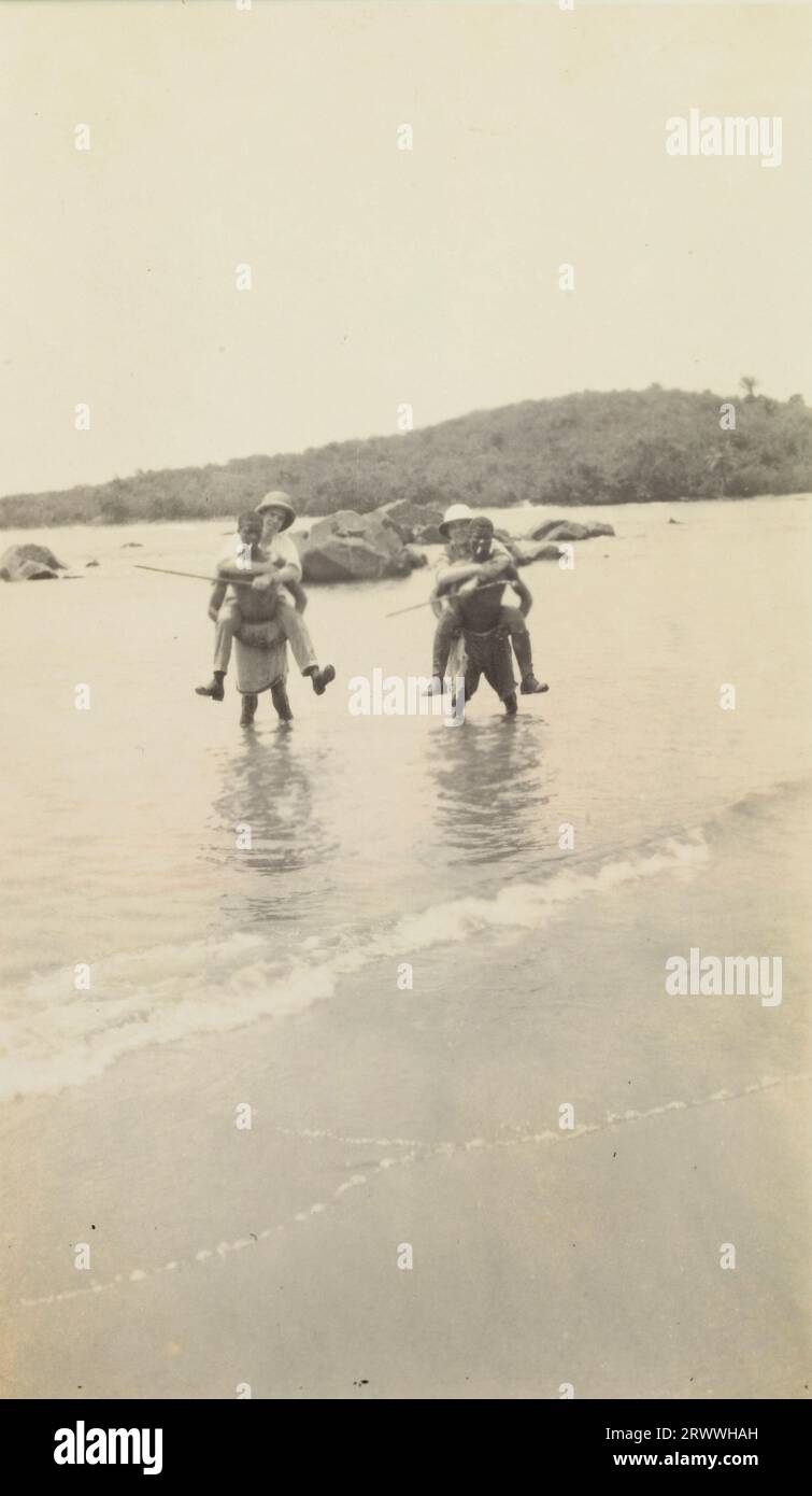 Alfred Tamlin and another European man, both wearing pith helmets, are carried in shallow waters on the backs of two African men who are smoking cigarettes. They are close to the beach and trees are visible on the hills behind them. Stock Photo