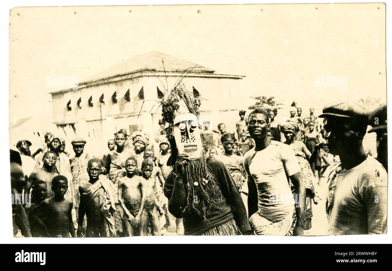 A crowd of African people form a throng in a town square in front of a large building. In the centre of the image is a man dressed in a costume with elaborate mask with foliage on top of it. Later manuscript caption: Witch Doctor. Stock Photo