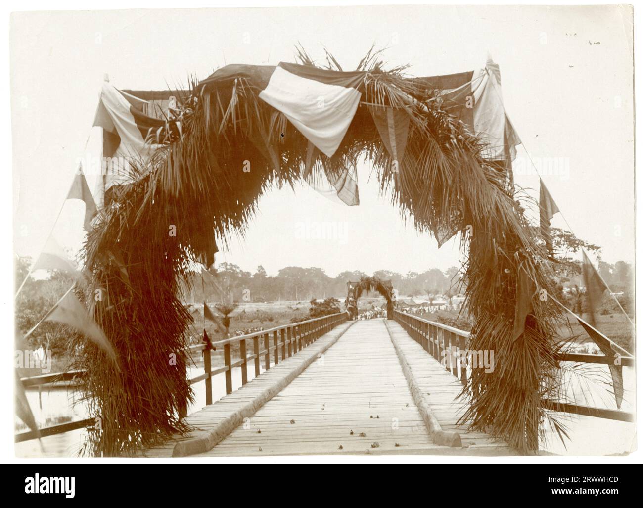 View across the Pusa Bridge in Nsawam, decorated ready for its opening by the Governor with flags and foliage strung up at either end. A large crowd of people are visible, congregated at the far side. Original manuscript caption: The new 'Pusa' bridge - opened by Sir Hugh Clifford - 1-7-18. Nsawam. Gold Coast. Stock Photo