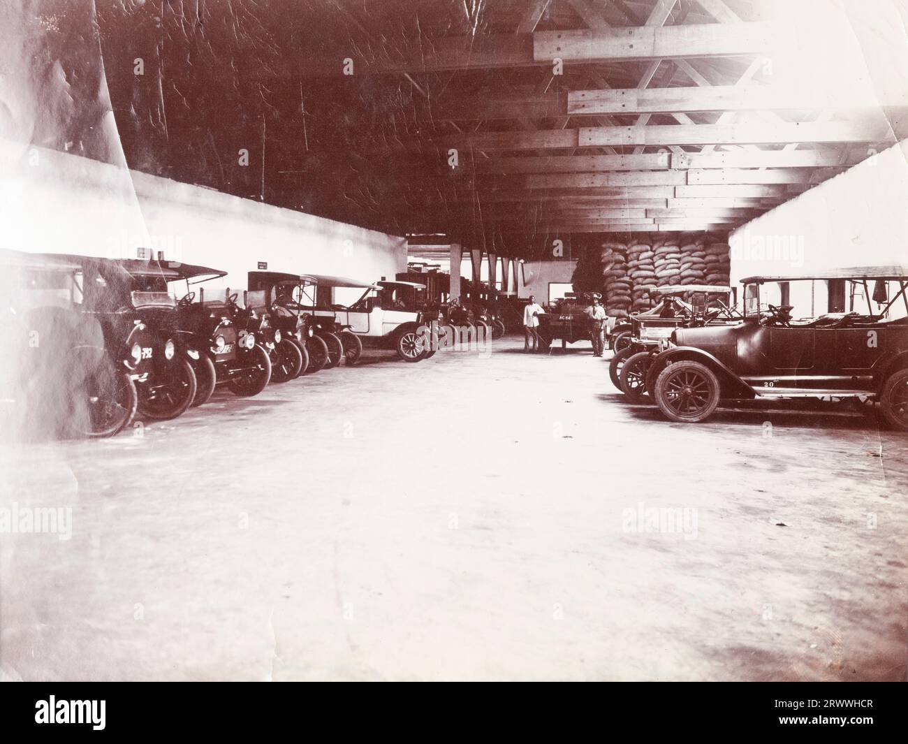 Interior of the garages of the mining company Swanzy & Co., showing around 12 cars neatly parked and a large pile of sandbags in the back corner. Two African men are standing with one of the cars. -Original manuscript caption: 4/4/18. Swanzy's Motor Transport Dep., Accra. Later manuscript caption: Motor Transport Depo Accra. Stock Photo