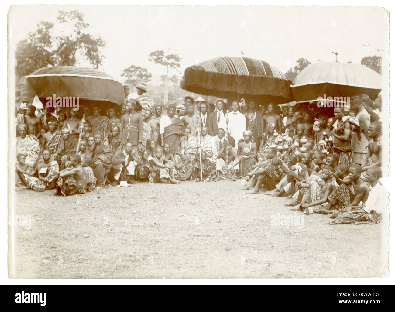 A large crowd of African men gather on an expanse of grass. They are grouped in a semicircle and some are sheltering under three large umbrellas. Some are dressed in traditional robes, others in Western-style office dress and some in police-style uniform. Original manuscript caption: Native chiefs 'Palaver'. Stock Photo