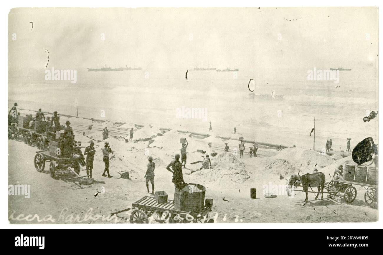 A busy scene on the beach at Accra with a large number of African labourers digging in heaps of sand and waggons being loaded up with boxes. At the water's edge a large pipeline is visible, and there are large ships out to sea in the distance. The caption to another image taken at the same location describes it as outside Swanzy's Motor Transport Dept. Captioned in the negative: Accra harbour [..26] 1919. Stock Photo