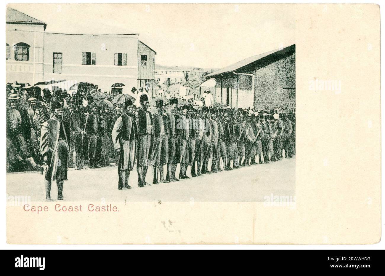 Postcard view of rows of African men lined up outside the castle at Cape Coast. The front row are in dress uniforms, possibly military, and those behind them in smart attire. There is a large crowd in the background. Printed caption: Cape Coast Castle. Stock Photo