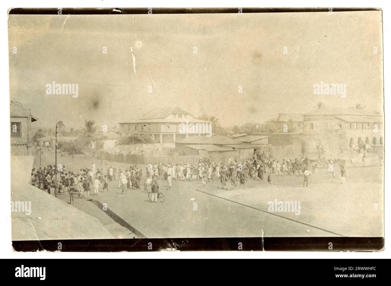 Street scene in Accra, showing a large crowd of people possibly waiting for an event or queuing to gain entry to something. The queue stretches across the street and includes people with bicycles and muscial instruments. Behind them are some grand buildings, with corrugated iron shacks nearer to the foreground. Original manuscript caption: Accra - Gold Coast. Stock Photo