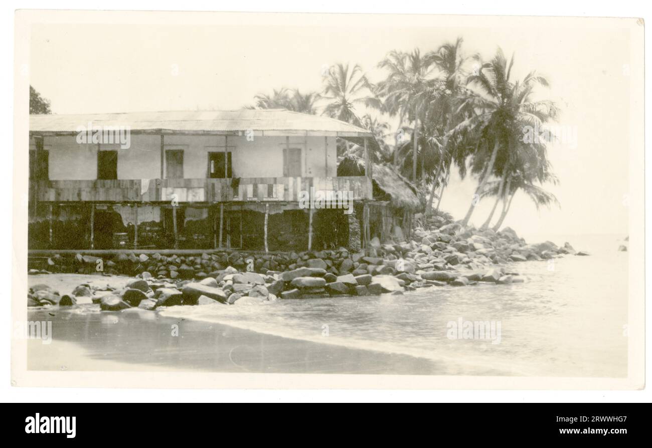 Exterior view of a large house on stilts built at the water's edge near a rocky cove, in an unidentified location. There is a first floor veranda and palm trees surrounding it. Stock Photo