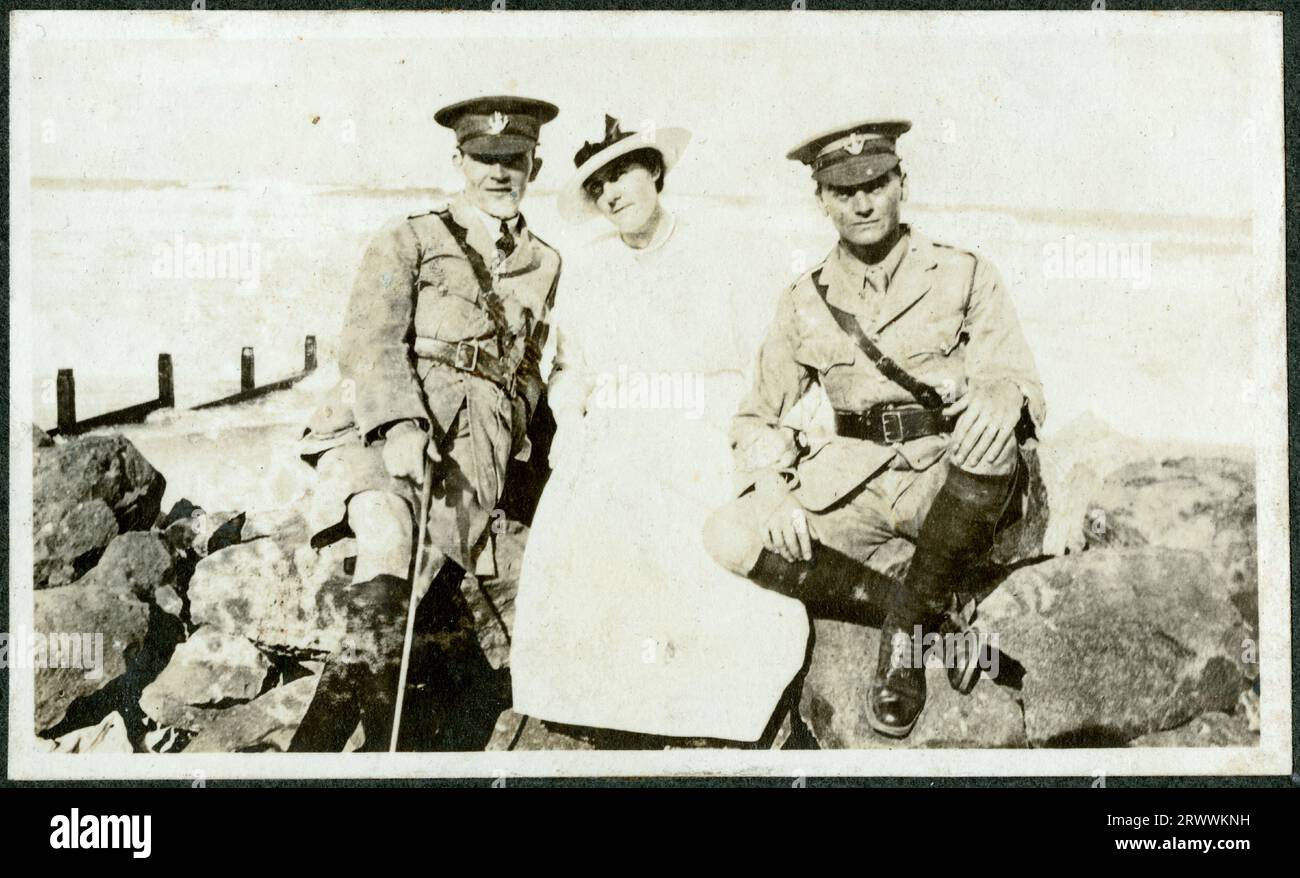 Two European officers sit either side of a smartly dressed woman on rocks next to the sea in Durban, with waves visible behind. One of the men is Bertie Rand.  Original manuscript caption: Along the beach at Durban. Stock Photo