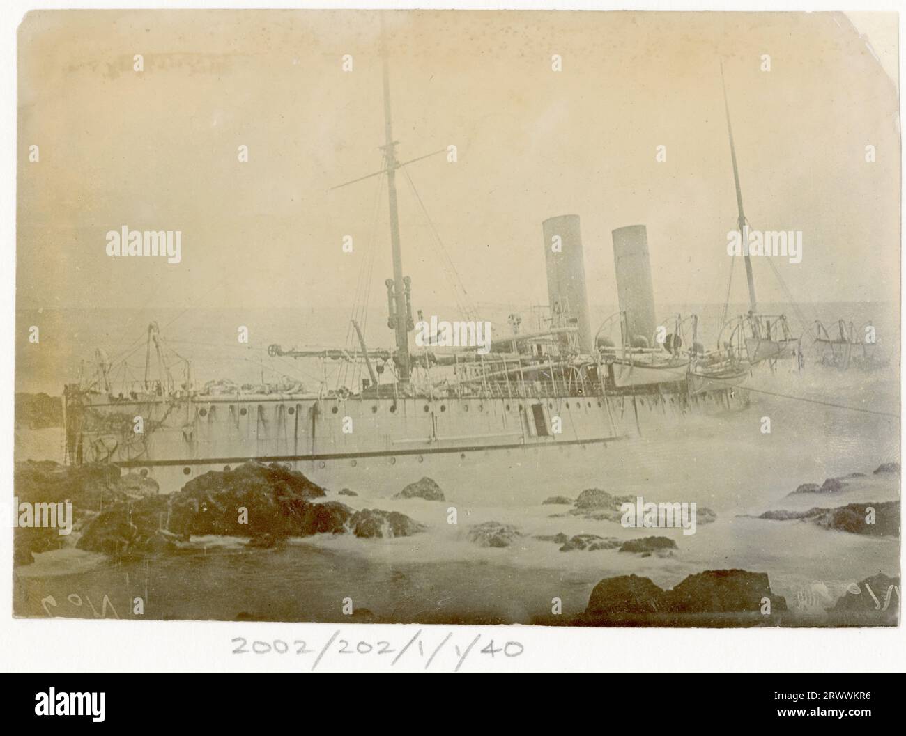 View of a two-funnelled, two-masted ship wrecked on coastal rocks at Mauritius. While the bow is mainly above water the stern section is submerged. Handwritten caption reads: Wreck of R.I.M.S. Warren Hastings. Stock Photo