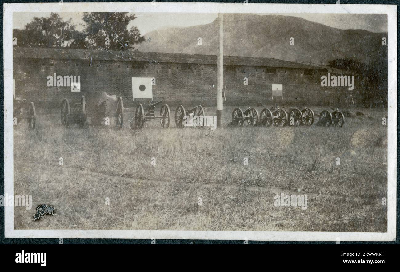 A line of artillery guns of varying sizes, resting on gun carriages, are lined up on grass next to the high wall of a building at the military camp in Zomba. Original manuscript caption: A few captured guns outside the boma at Zomba, Central Africa. Stock Photo