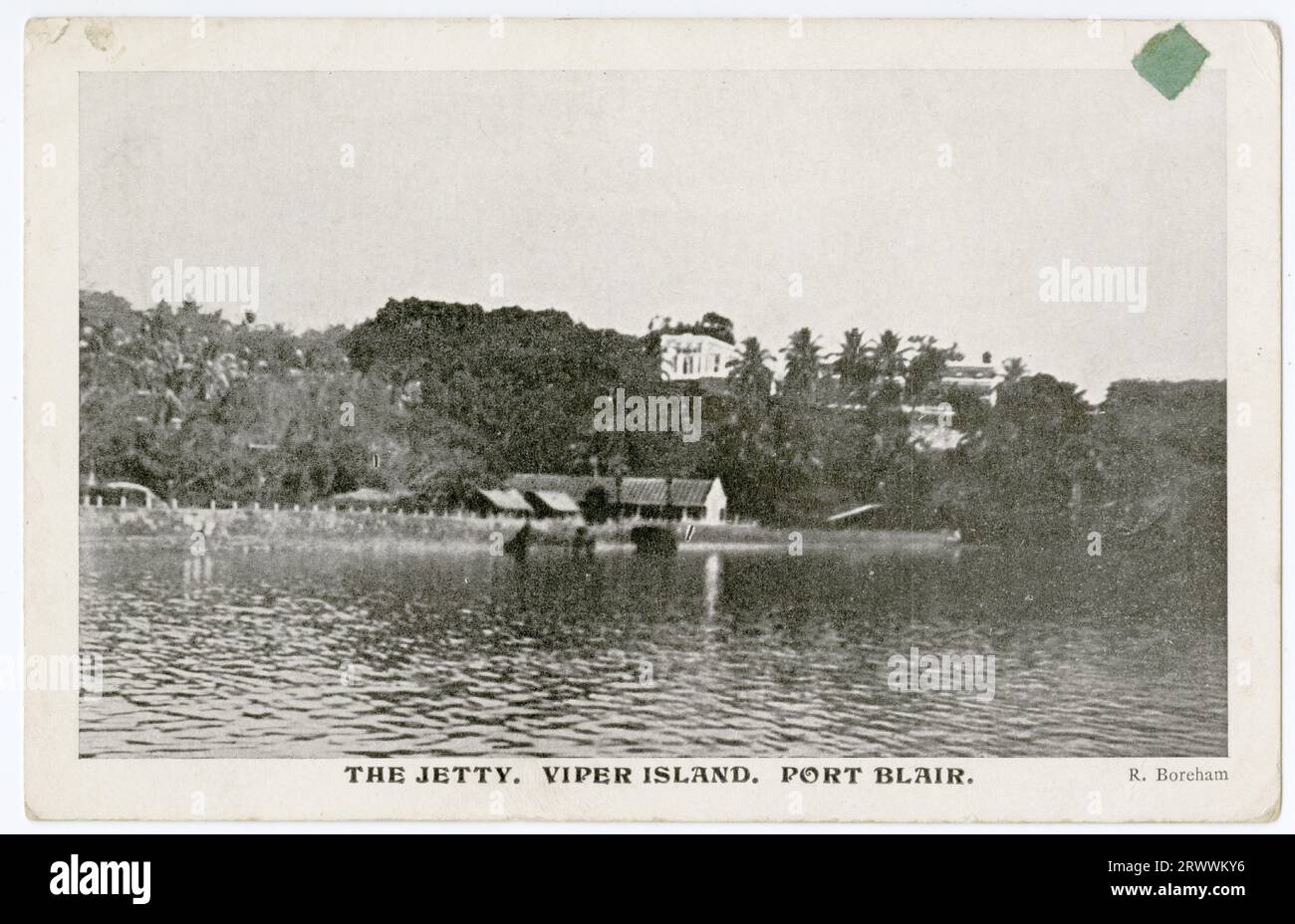 Postcard showing a small group of sheds on a waterfront below a tree-clad hill, on which is sited a substantial building, partly hidden among the trees. A printed caption reads The Jetty, Viper Island, Port Blair. Copyright: R Boreham. Stock Photo