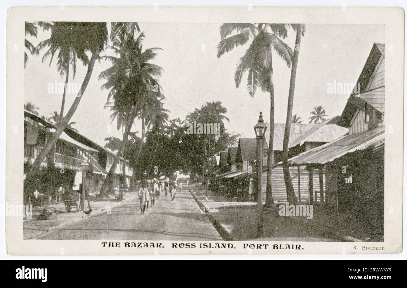 Postcard showing a palm tree-lined street, with two storey wooden framed buildings on each side. The buildings have upper verandahs on one side and upper gables on the other. A line of pedestrians is shown walking along the centre of the road. Described in printed caption as The Bazaar, Ross Island, Port Blair. Caption on reverse reads: Native Shops. My back verandah looks down this street. Copyright: R Boreham. Stock Photo