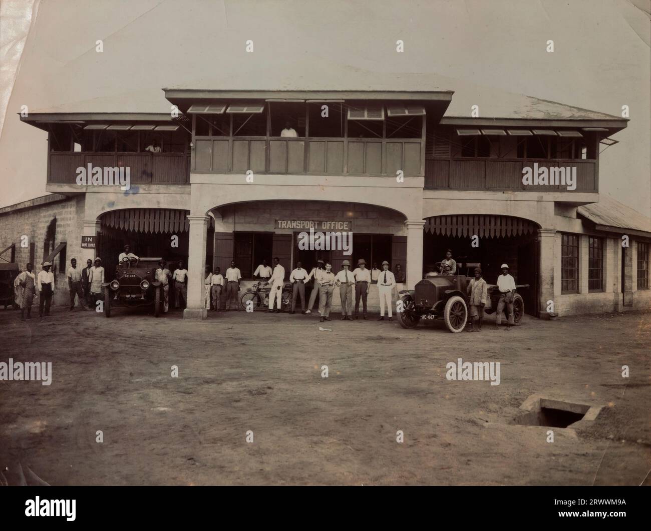 Front elevation of the transport department of the mining company Swanzy & Co. Employees pose for a group photograph outside a large stone building signed 'Transport Office' with garages underneath and offices above. There are two cars and a motorbike parked outside the garages. Four of the employees, including Mr Tamlin, are European; the rest are African. The majority are smartly dressed with a few in mechanics' clothes. Stock Photo