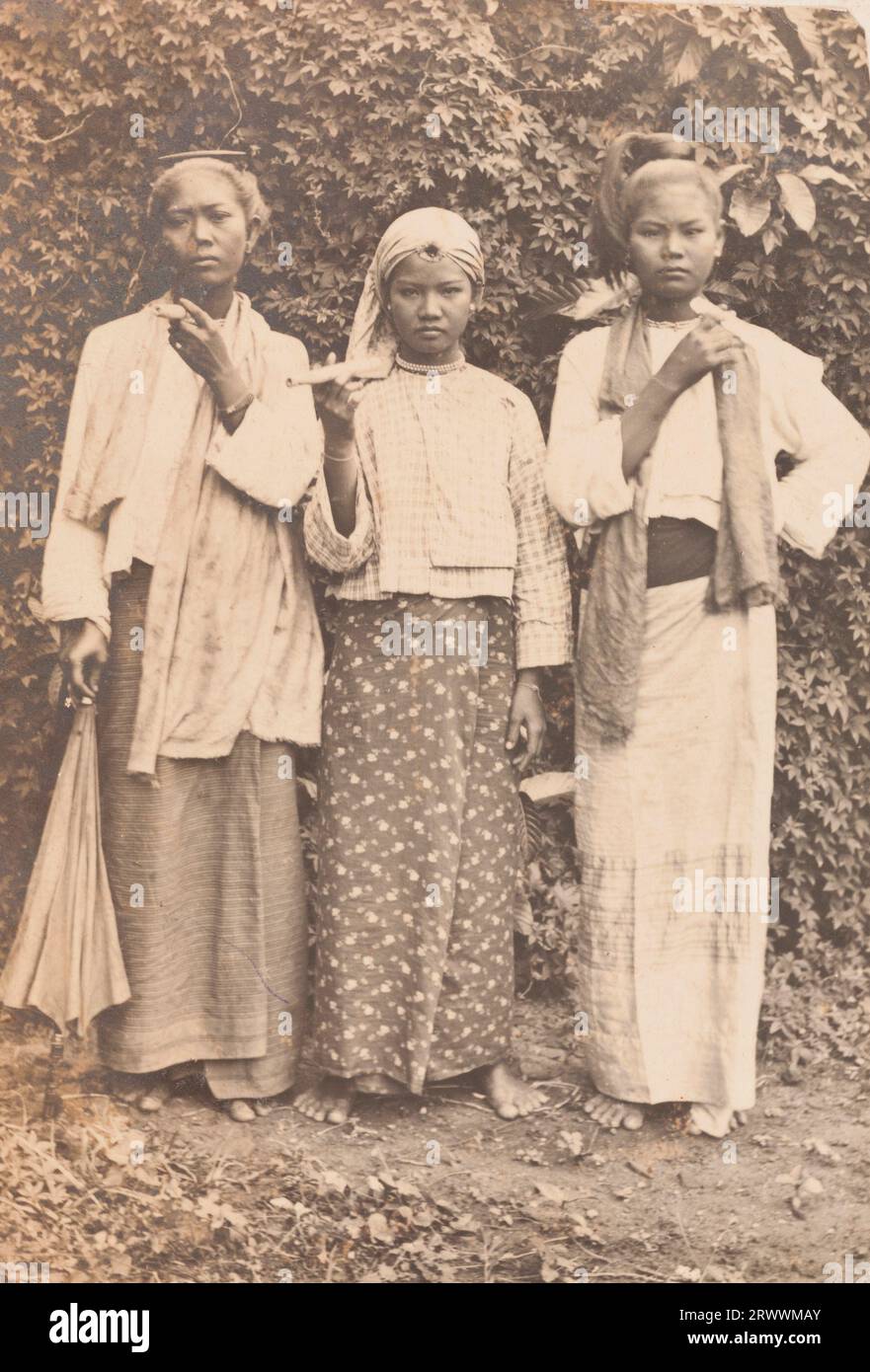 Portrait of three Burmese woman in traditional dress including a silk longyi (wrapped cloth skirt) holding pipes. They stand barefoot outside, in front of foliage. Stock Photo
