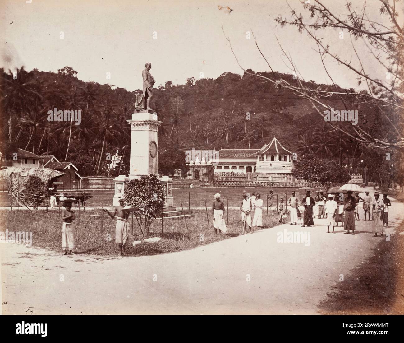 Bronze statue of Sir Henry Ward stands on a tall plinth in front of the Sri Dalada Maligawa, Temple of the Sacred Tooth Relic. A group of local men stand in the foreground, some shaded by umbrellas, and there is an ox-drawn cart beside the statue. Caption reads: Kandy Sir Henry Ware Temple containing Buddha's Tooth. Stock Photo