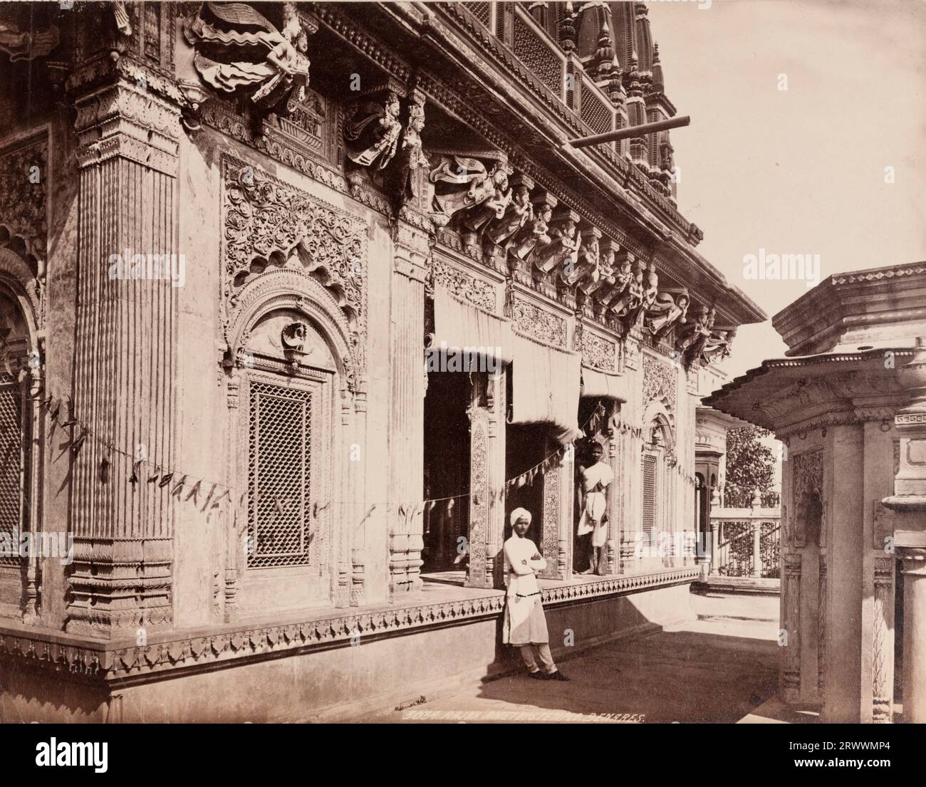 View of the Amethi Shiva Temple on the Manikarnika Ghat in Benares [Varanasi], built by the King of Amethi, Raja Lal Madhav Singh. The exterior walls of the temple are intricately carved and winged figures on the brackets supporting the second floor can be seen. A string of dry mango leaves is hung outside the entrance. Two Indian men in white traditional dress rest against the walls outside. Inscribed on negative: Frith's Series. 3034 Rajah Amethi's Temple Benares. Caption reads: Rajah Amethi's Temple, Benares. Stock Photo