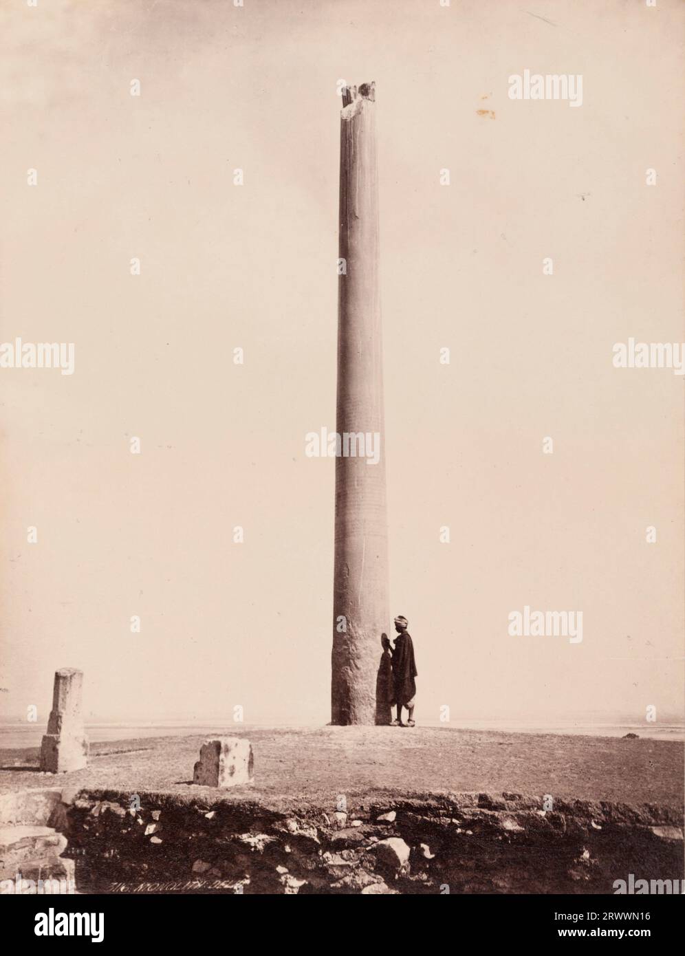 View of the Ashoka pillar with its broken top and inscriptions. An Indian person in traditional dress stands at the base of the Buddhist monument, with one hand against the stone. Inscribed on negative: Frith's Series. 3116. Monolith: Delhi. Caption reads: Asoka's Pillar, (B.C. 200) Delhi. Stock Photo
