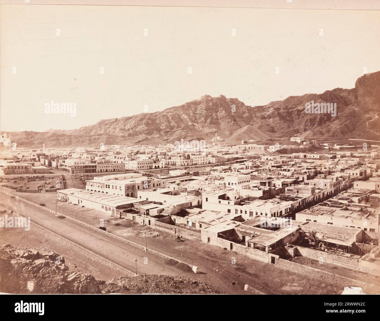 View of Aden, taken from a high vantage point with rocky mountains in the background. The buildings within the city are laid out in a grid-like formation with a fenced strip of land where animals are grazing. Caption reads: Aden. Stock Photo