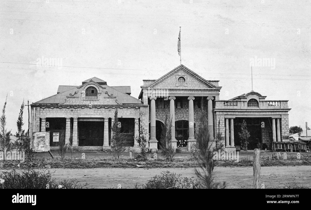 Front elevation of the Nairobi Assembly Rooms, showing the facade of the building. The exterior looks messy, with overgrown shrubs and broken fences. A poster advertises a boxing event and there is a plaque on one part of the building for P.A. Raphael, Auctioneer. Original manuscript caption: Old Palace Theatre 1912. 6th Avenue. Stock Photo